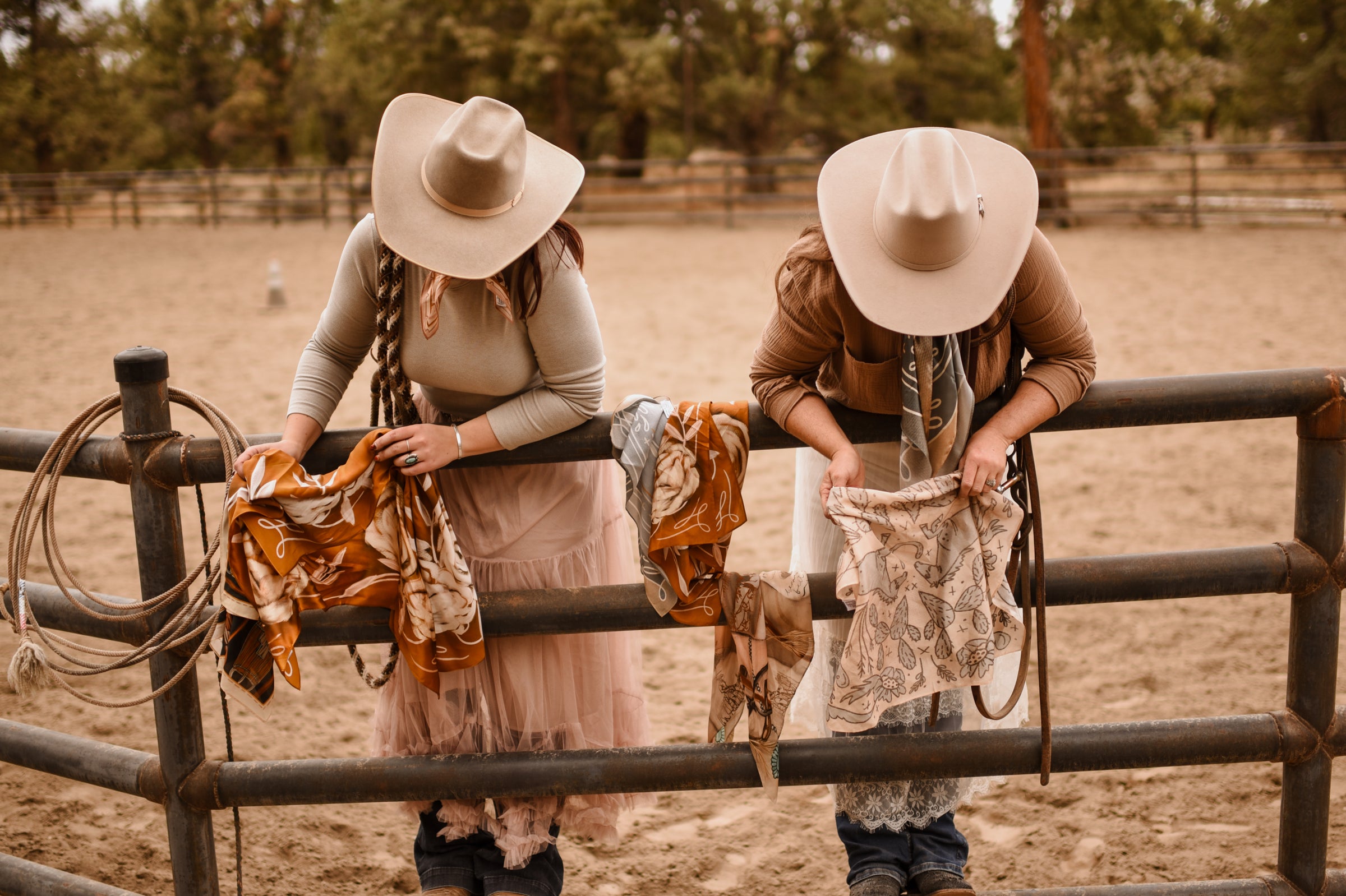 Two people in cowboy hats standing behind a fence with lassos and patterned wild rags in a rural setting.